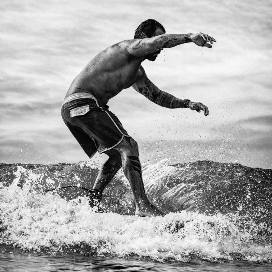 Rapha Preto a surfar, de braços abertos sobre a espuma da onda — fotografia a preto e branco